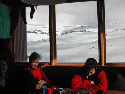 Two adults sitting inside Illawong Lodge, with a snow-covered mountain range through the window behind them